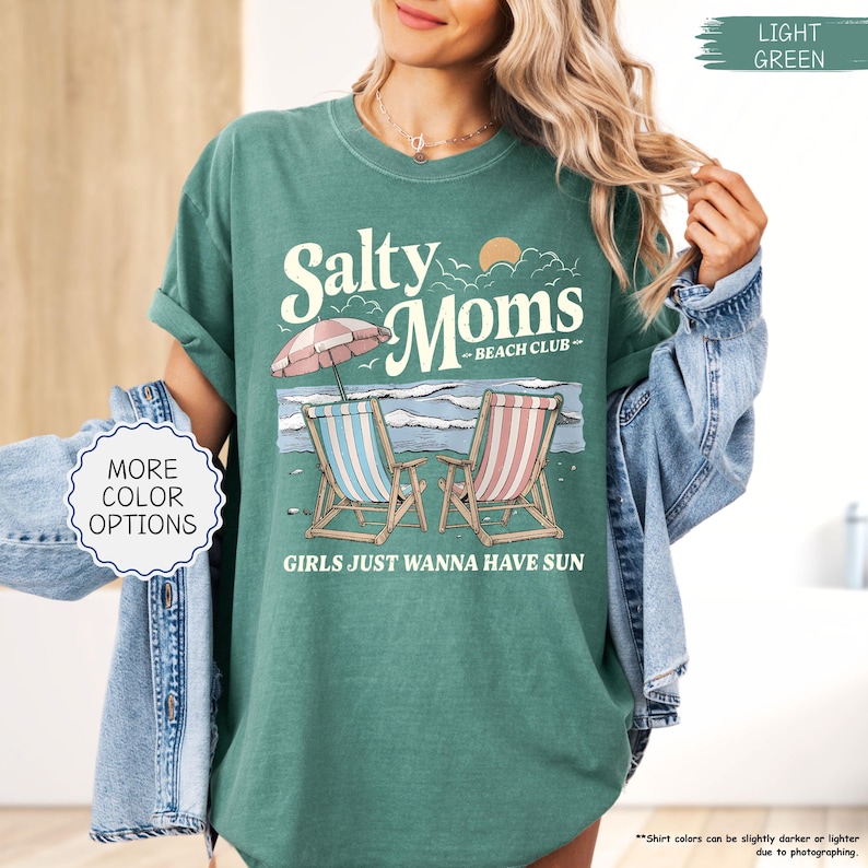 May include: A light green t-shirt with the text "Salty Moms Beach Club" and the phrase "Girls Just Wanna Have Sun." The graphic includes beach chairs, an umbrella, and ocean waves. The shirt has a relaxed fit.