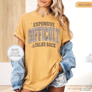 May include: Mustard-colored t-shirt with the text "EXPENSIVE DIFFICULT & TALKS BACK" in a distressed, leopard print font. The shirt has rolled-up sleeves and a relaxed fit. The image also shows a denim jacket.