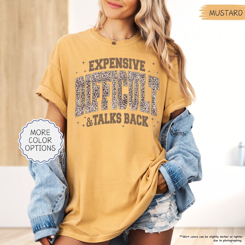 May include: Mustard-colored t-shirt with the text "EXPENSIVE DIFFICULT & TALKS BACK" in a distressed, leopard print font. The shirt has rolled-up sleeves and a relaxed fit. The image also shows a denim jacket.