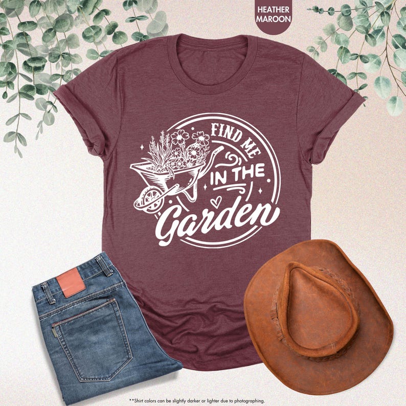 Puede incluir: Una camiseta de color marr&oacute;n rojizo con un gr&aacute;fico blanco de una carretilla llena de flores y el texto "Find me in the Garden".