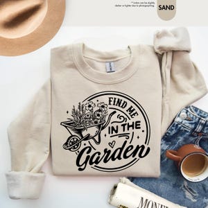 Puede incluir: Una sudadera de color crema con un dise&ntilde;o gr&aacute;fico negro de una carretilla llena de flores y el texto "FIND ME IN THE GARDEN".