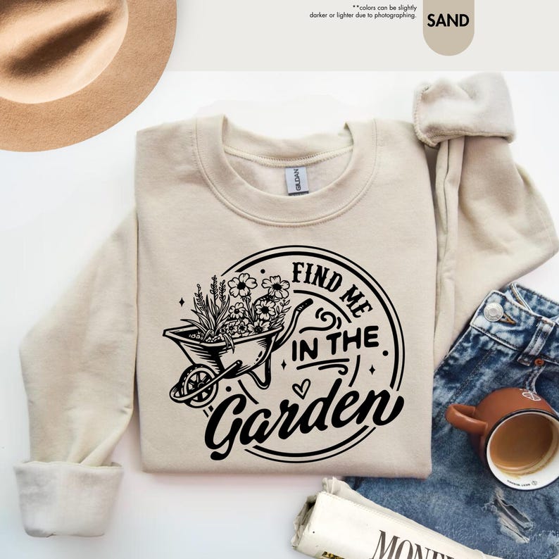 Puede incluir: Una sudadera de color crema con un dise&ntilde;o gr&aacute;fico negro de una carretilla llena de flores y el texto "FIND ME IN THE GARDEN".