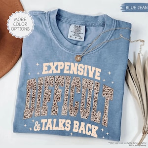 May include: A blue jean colored t-shirt with the words "EXPENSIVE, DIFFICULT, & TALKS BACK" in a tan font with a leopard print design. The shirt has a crew neck and short sleeves. A gold necklace is draped across the shirt.