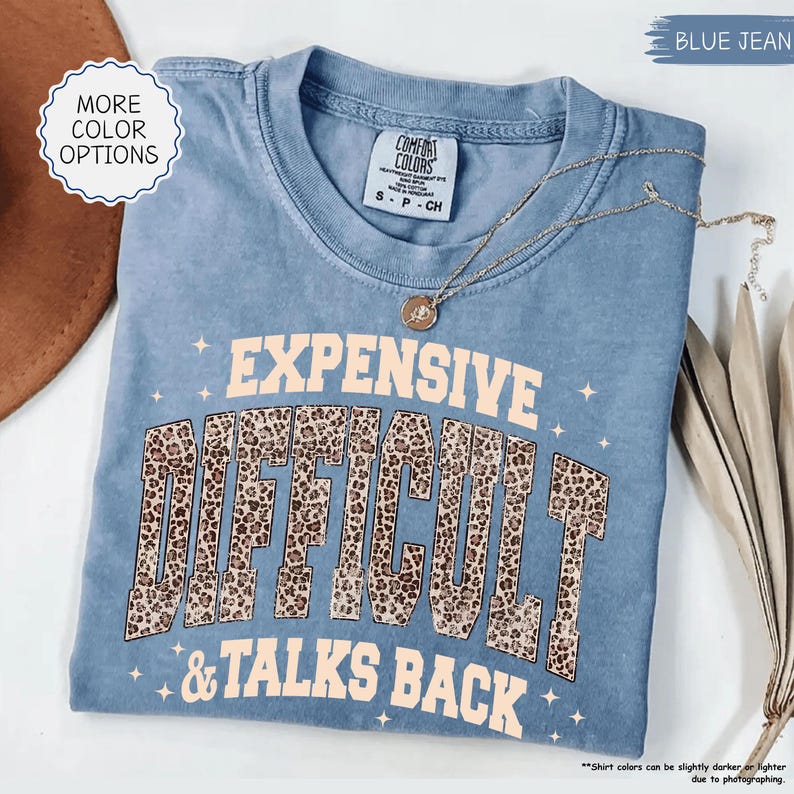 May include: A blue jean colored t-shirt with the words "EXPENSIVE, DIFFICULT, & TALKS BACK" in a tan font with a leopard print design. The shirt has a crew neck and short sleeves. A gold necklace is draped across the shirt.