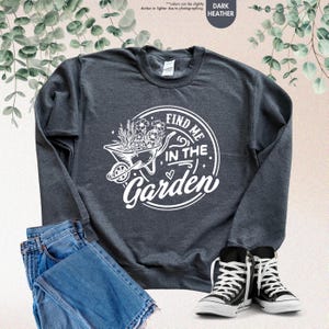 Puede incluir: Sudadera gris oscuro jaspeada con un gr&aacute;fico blanco de una carretilla llena de flores y el texto "Find me in the garden".