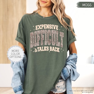 May include: Moss green t-shirt with the text "EXPENSIVE, DIFFICULT, & TALKS BACK" in a white and leopard print design. The shirt has rolled-up sleeves and is paired with a denim jacket. The image also includes a "MORE COLOR OPTIONS" button.