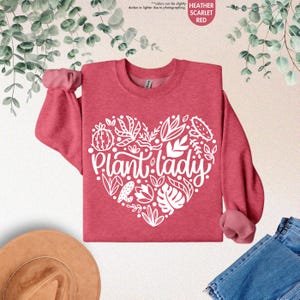 May include: A heather scarlet red sweatshirt with a white heart-shaped design featuring the text "Plant lady" surrounded by various plant illustrations.