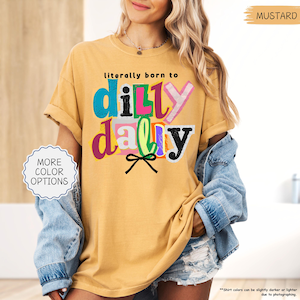 K&ouml;nnte beinhalten: Senffarbenes T-Shirt mit dem Text "literally born to dilly dally" in bunten, verspielten Buchstaben. Das Shirt hat hochgekrempelte &Auml;rmel und eine lockere Passform. Ein Knopf auf der linken Seite zeigt "More Color Options".