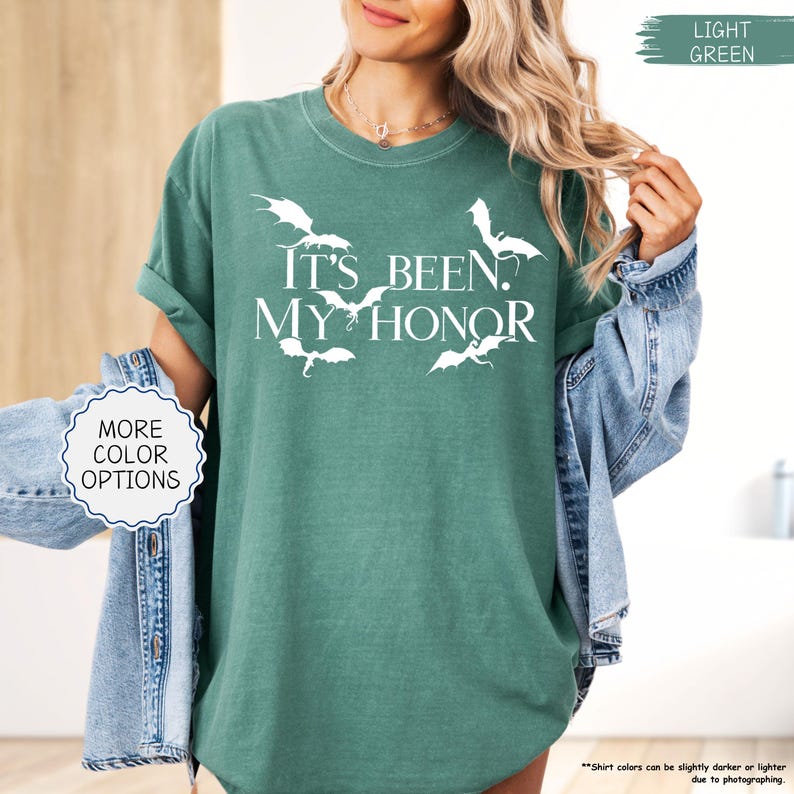 Pu&ograve; includere: T-shirt verde chiaro con la scritta bianca "IT'S BEEN. MY HONOR" e grafiche di pipistrelli bianchi. La maglietta ha maniche corte e girocollo. &Egrave; visibile anche un cerchio bianco con "MORE COLOR OPTIONS".