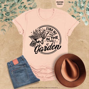 Puede incluir: Una camiseta rosa con un gr&aacute;fico negro de una carretilla llena de flores y el texto "Find me in the Garden".
