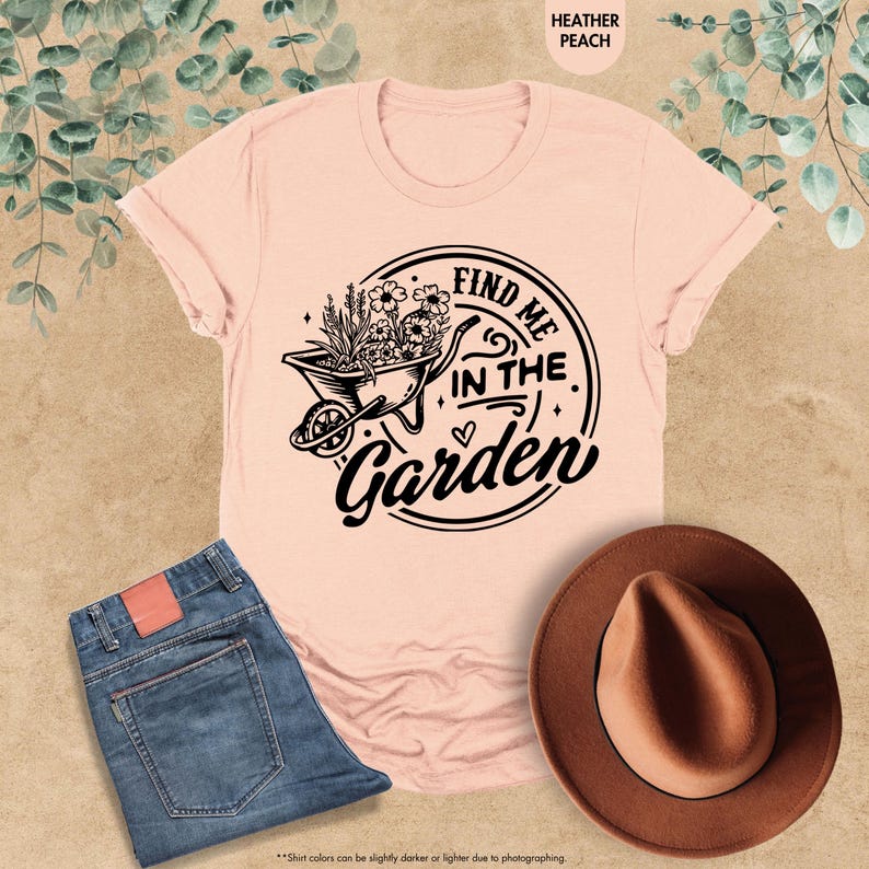 Puede incluir: Una camiseta rosa con un gr&aacute;fico negro de una carretilla llena de flores y el texto "Find me in the Garden".