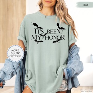Pu&ograve; includere: T-shirt verde chiaro con la scritta nera "It's Been My Honor", accompagnata da sagome di pipistrelli neri. La maglietta ha le maniche arrotolate e fa parte di una collezione con pi&ugrave; opzioni di colore.