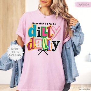 K&ouml;nnte beinhalten: Hellrosa T-Shirt mit dem Schriftzug "literally born to dilly dally" in bunten Buchstaben. Das Shirt hat eine lockere Passform und kurze &Auml;rmel. Eine Jeansjacke ist &uuml;ber den Schultern drapiert. Die Worte "More Color Options" stehen in einem wei&szlig;en Kreis.