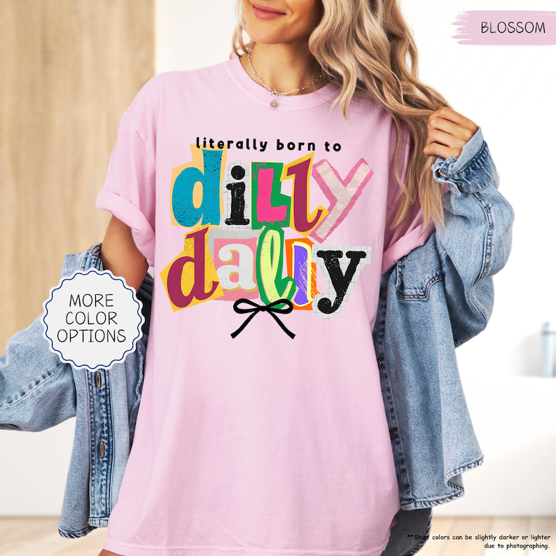 K&ouml;nnte beinhalten: Hellrosa T-Shirt mit dem Schriftzug "literally born to dilly dally" in bunten Buchstaben. Das Shirt hat eine lockere Passform und kurze &Auml;rmel. Eine Jeansjacke ist &uuml;ber den Schultern drapiert. Die Worte "More Color Options" stehen in einem wei&szlig;en Kreis.