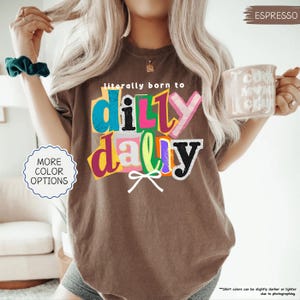 K&ouml;nnte beinhalten: Espressofarbenes T-Shirt mit dem Text "literally born to ditty dally" in bunten, geschichteten Buchstaben. Das Shirt hat eine lockere Passform und einen Rundhalsausschnitt. Am unteren Rand des Textes befindet sich eine wei&szlig;e Schleife. Teil einer Bekleidungskollektion.