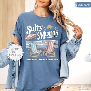 May include: Blue jean colored t-shirt with the text "Salty Moms Beach Club" and a beach scene graphic. The graphic includes two beach chairs, an umbrella, and the phrase "Girls Just Wanna Have Sun."