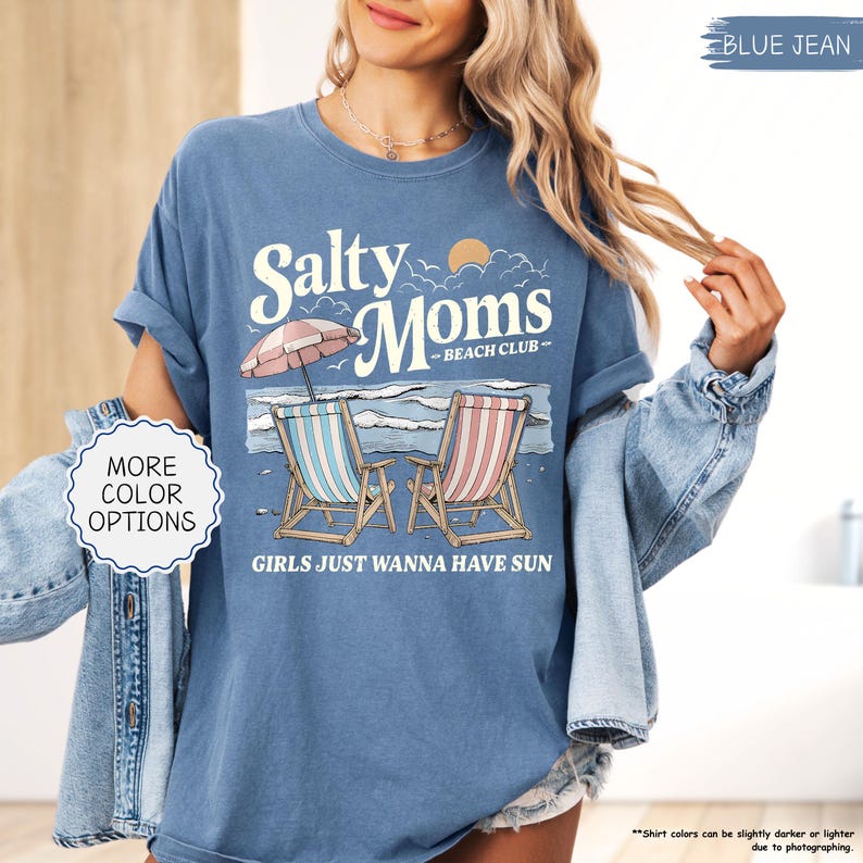 May include: Blue jean colored t-shirt with the text "Salty Moms Beach Club" and a beach scene graphic. The graphic includes two beach chairs, an umbrella, and the phrase "Girls Just Wanna Have Sun."