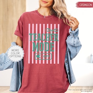 May include: Crimson-colored t-shirt with the text "TEACHER MODE OFF" in teal, set against a pink and white striped background. The shirt has a relaxed fit and rolled-up sleeves. The words "MORE COLOR OPTIONS" are in a white circle.