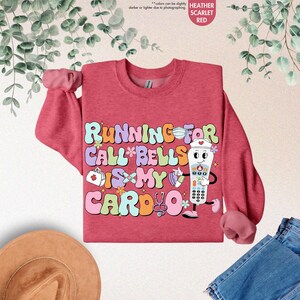 May include: A heather scarlet red sweatshirt with a graphic design of a cartoon nurse holding a call bell. The text on the sweatshirt reads "Running for call bells is my cardio".