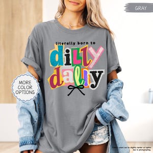 K&ouml;nnte beinhalten: Graues T-Shirt mit dem Schriftzug "literally born to dilly dally" in farbenfrohem, geschichtetem Text. Das Shirt hat hochgekrempelte &Auml;rmel und wird mit einer Jeansjacke kombiniert. Die Worte "MORE COLOR OPTIONS" stehen in einem wei&szlig;en Kreis.