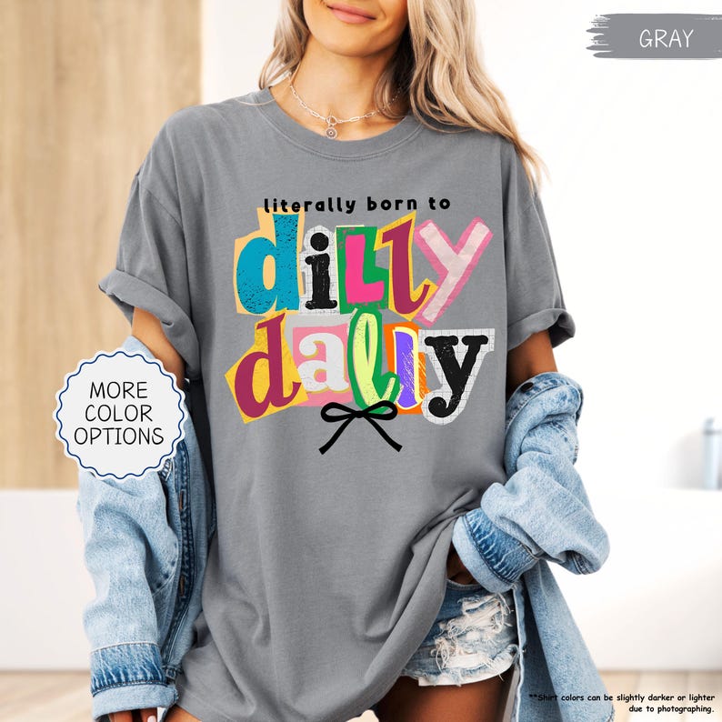 K&ouml;nnte beinhalten: Graues T-Shirt mit dem Schriftzug "literally born to dilly dally" in farbenfrohem, geschichtetem Text. Das Shirt hat hochgekrempelte &Auml;rmel und wird mit einer Jeansjacke kombiniert. Die Worte "MORE COLOR OPTIONS" stehen in einem wei&szlig;en Kreis.
