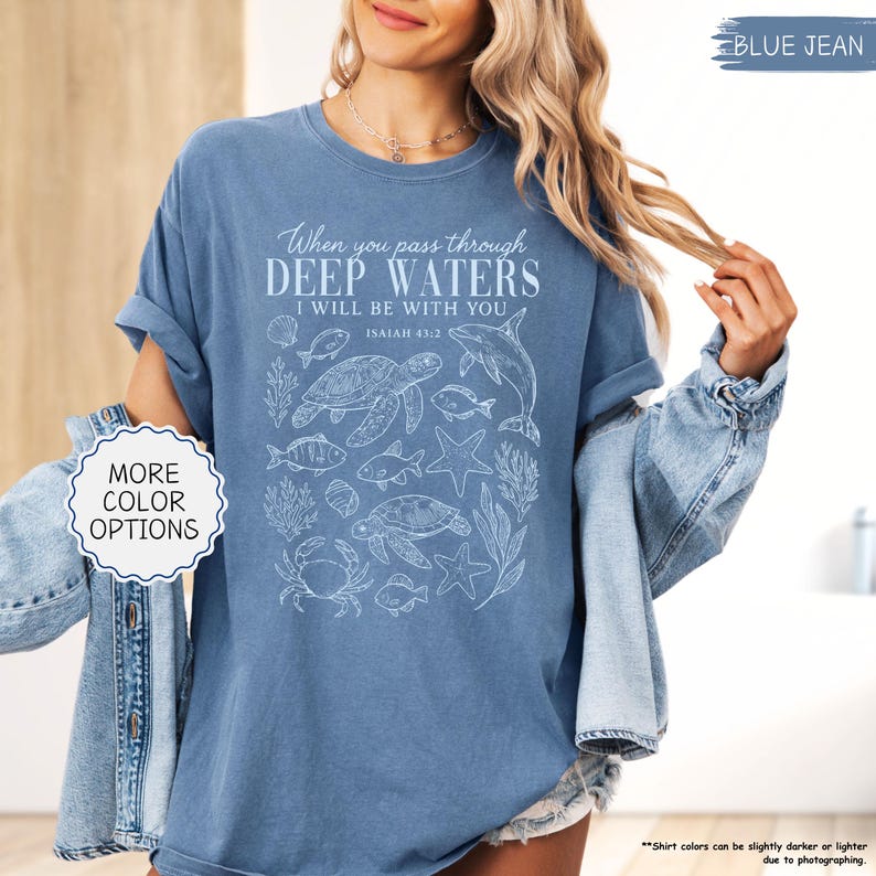 May include: Blue jean-colored t-shirt with the text "When you pass through deep waters I will be with you Isaiah 43:2". The shirt features a white graphic of sea creatures. The words "More Color Options" are in a white circle.