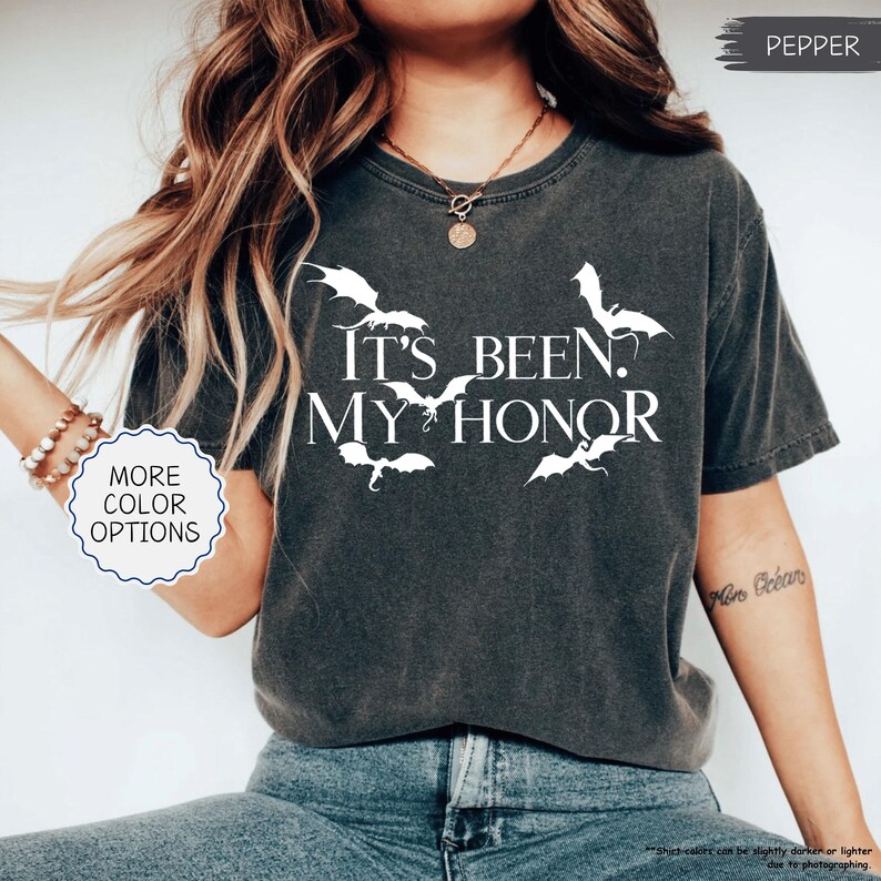 May include: A dark gray t-shirt with the text "IT'S BEEN. MY HONOR" in white, along with white bat graphics. The shirt has a vintage, worn look. The words "MORE COLOR OPTIONS" are also visible.