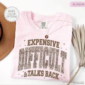 May include: A light pink Comfort Colors t-shirt with the words "EXPENSIVE DIFFICULT & TALKS BACK" in a brown leopard print font. A delicate necklace with a pendant rests on the shirt. The shirt has a round neckline.