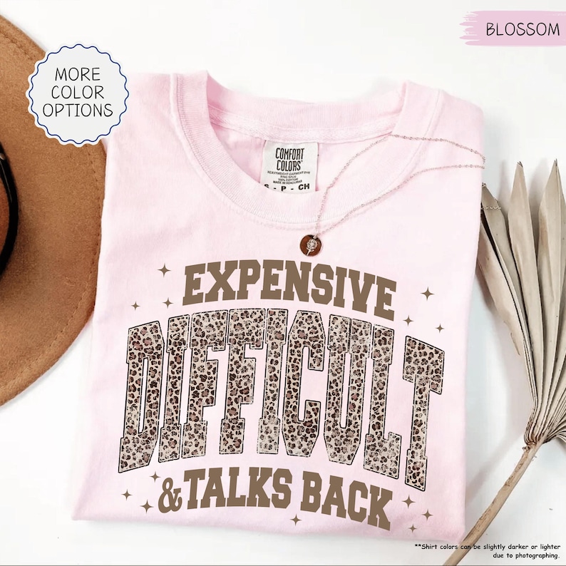 May include: A light pink Comfort Colors t-shirt with the words "EXPENSIVE DIFFICULT & TALKS BACK" in a brown leopard print font. A delicate necklace with a pendant rests on the shirt. The shirt has a round neckline.