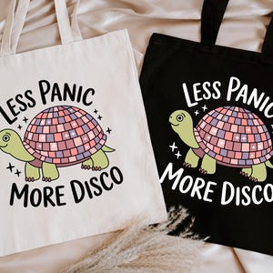 May include: Two tote bags, one cream and one black, each featuring a cartoon turtle with a disco ball shell. The text "Less Panic More Disco" is above and below the turtle illustration. The bags have long handles.
