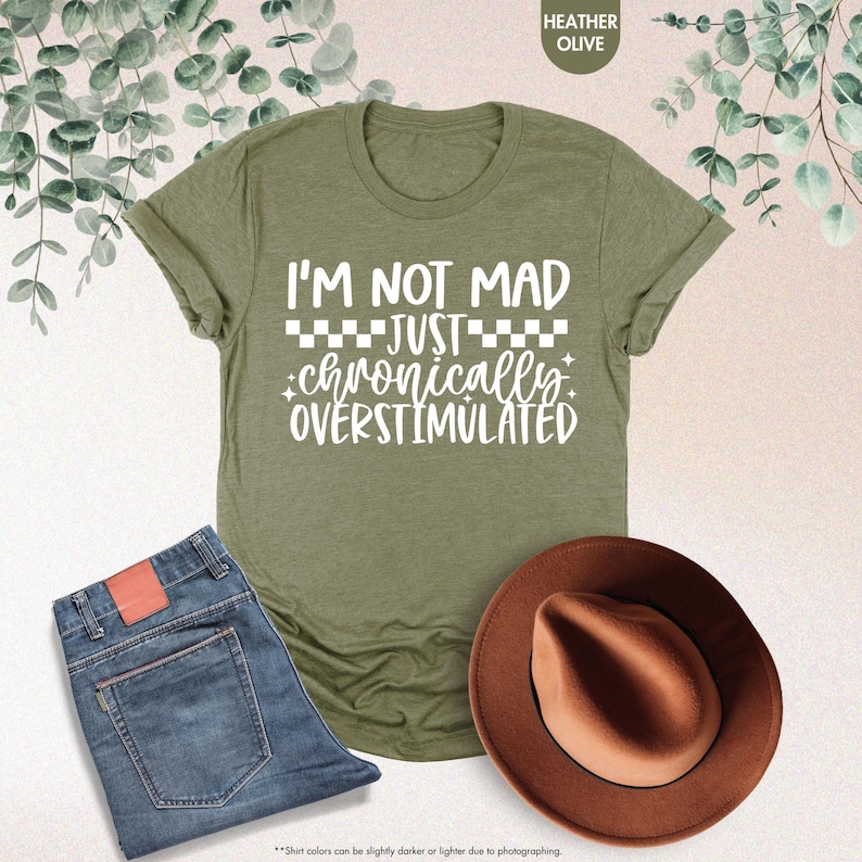 Op de afbeelding: Een olijfgroen t-shirt met de tekst "I'M NOT MAD JUST chronically OVERSTIMULATED" in het wit. Het shirt wordt gecombineerd met een blauwe jeans en een bruine hoed. De afbeelding bevat ook een takje groen.