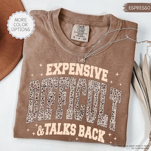 May include: Espresso-colored t-shirt with the words "EXPENSIVE DIFFICULT & TALKS BACK" in a tan font with leopard print accents. The shirt has a crew neck and short sleeves. A silver necklace with a pendant is draped across the shirt.