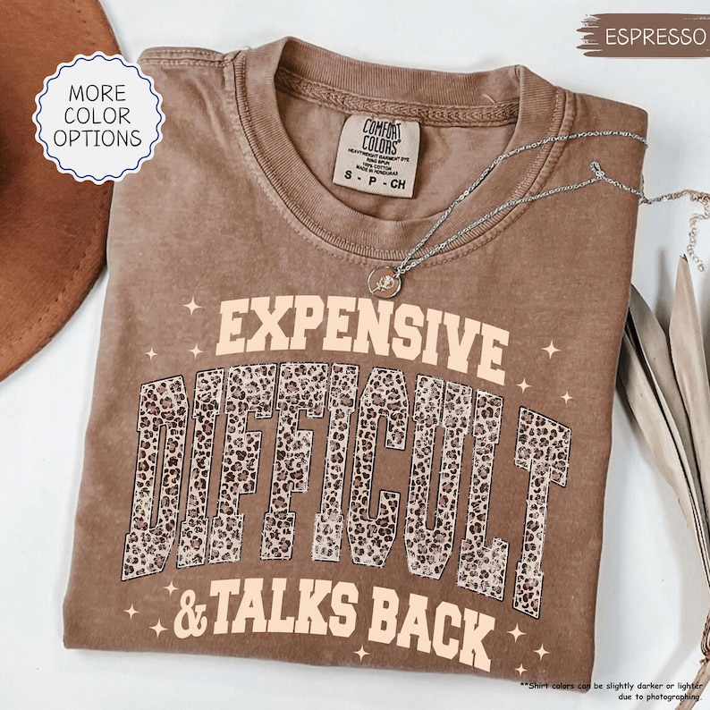 May include: Espresso-colored t-shirt with the words "EXPENSIVE DIFFICULT & TALKS BACK" in a tan font with leopard print accents. The shirt has a crew neck and short sleeves. A silver necklace with a pendant is draped across the shirt.
