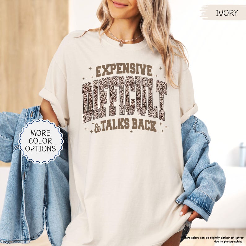 May include: Ivory-colored t-shirt with the text "EXPENSIVE DIFFICULT & TALKS BACK" in a brown leopard print design. The shirt has rolled-up sleeves and is paired with a denim jacket. The words "MORE COLOR OPTIONS" are also visible.