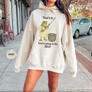 Pu&ograve; includere: Felpa con cappuccio color sabbia con un'illustrazione di una rana che tiene un cucchiaio sopra una pentola di zuppa. Il testo sulla felpa dice "That's it. You're going in the SOUP."