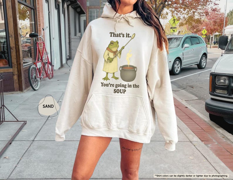 Pu&ograve; includere: Felpa con cappuccio color sabbia con un'illustrazione di una rana che tiene un cucchiaio sopra una pentola di zuppa. Il testo sulla felpa dice "That's it. You're going in the SOUP."