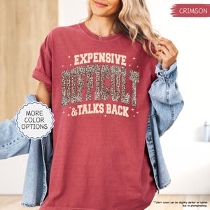May include: Crimson-colored t-shirt with the text "EXPENSIVE DIFFICULT & TALKS BACK" in a bold, stylized font. The word "DIFFICULT" features a leopard print pattern. The shirt has a vintage, worn look.