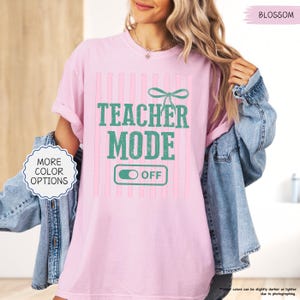 May include: A light pink t-shirt with the words "TEACHER MODE OFF" in teal lettering. The design includes vertical pink stripes and a teal bow. The shirt also has a "MORE COLOR OPTIONS" button.