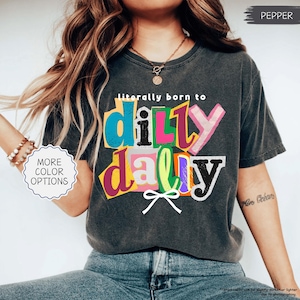 K&ouml;nnte beinhalten: Dunkelgraues T-Shirt mit dem Schriftzug "literally born to dilly dally" in bunten, ausgeschnittenen Buchstaben. Das Shirt hat einen Vintage-Look und eine kleine Schleifen-Grafik.