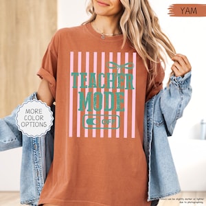 May include: A yam-colored t-shirt with a vertical pink and white striped design. The shirt features the text "TEACHER MODE OFF" in teal. A button on the shirt says "OFF". The shirt is a casual style.