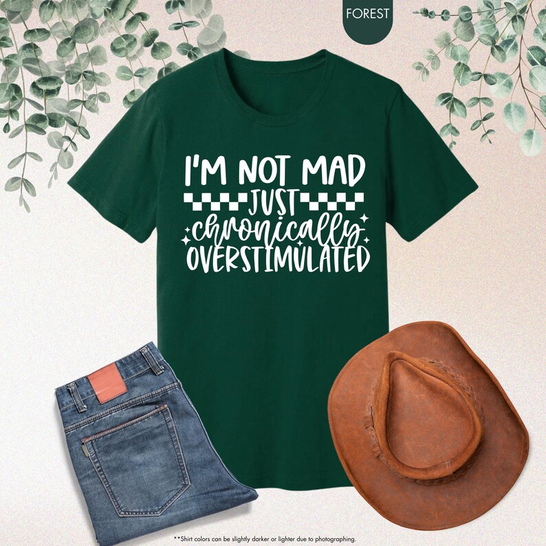 Op de afbeelding: Een donkergroen t-shirt met de tekst "I'M NOT MAD JUST chronically OVERSTIMULATED" in het wit. De afbeelding bevat ook een spijkerbroek en een bruine cowboyhoed.