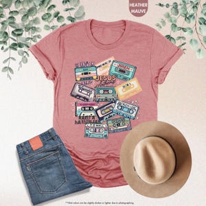 May include: A heather mauve t-shirt with a graphic design of vintage cassette tapes featuring Christian music titles like "Waymaker", "Reckless Love", "Oceans", and "It Is Well". The text "Jesus Anthems" is also included in the design.