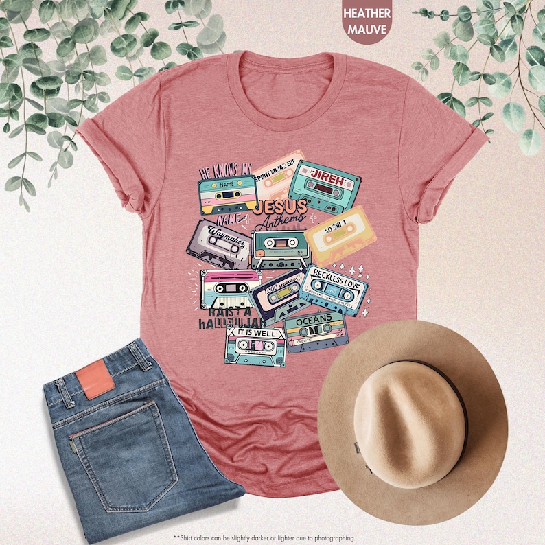 May include: A heather mauve t-shirt with a graphic design of vintage cassette tapes featuring Christian music titles like "Waymaker", "Reckless Love", "Oceans", and "It Is Well". The text "Jesus Anthems" is also included in the design.