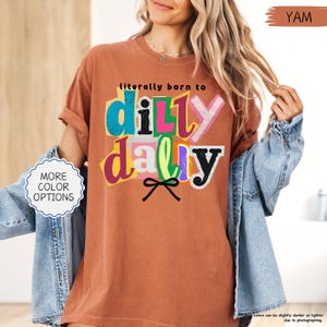K&ouml;nnte beinhalten: Ein terrakottafarbenes T-Shirt mit dem Schriftzug "literally born to dilly dally" in einem farbenfrohen, ausgeschnittenen Stil. Das Shirt hat hochgekrempelte &Auml;rmel und wird mit einer Jeansjacke kombiniert. Der Text "MORE COLOR OPTIONS" ist ebenfalls sichtbar.