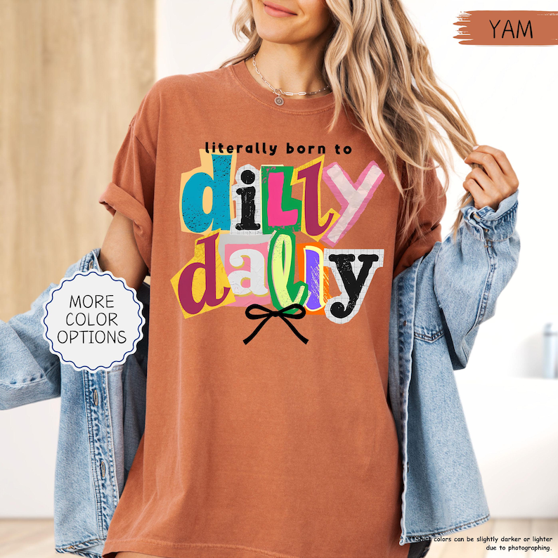 K&ouml;nnte beinhalten: Ein terrakottafarbenes T-Shirt mit dem Schriftzug "literally born to dilly dally" in einem farbenfrohen, ausgeschnittenen Stil. Das Shirt hat hochgekrempelte &Auml;rmel und wird mit einer Jeansjacke kombiniert. Der Text "MORE COLOR OPTIONS" ist ebenfalls sichtbar.