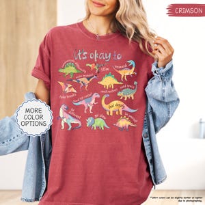 Puede incluir: Camiseta color carmes&iacute; con la frase "It's okay to" e ilustraciones de dinosaurios. La camiseta presenta frases como "communicate differently", "make mistakes" y "be yourself". Un bot&oacute;n a la izquierda dice "MORE COLOR OPTIONS".