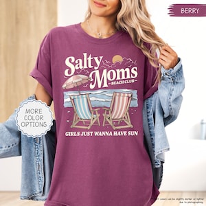May include: Berry-colored t-shirt with the text "Salty Moms Beach Club" and a beach scene graphic. The graphic includes two beach chairs, an umbrella, and the phrase "Girls Just Wanna Have Sun."