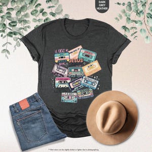 May include: A dark heather grey t-shirt with a design of vintage cassette tapes featuring Christian song titles like "He Knows My Name", "Waymaker", "Jireh", "It Is Well", "Reckless Love", "Oceans", and "So Will I".