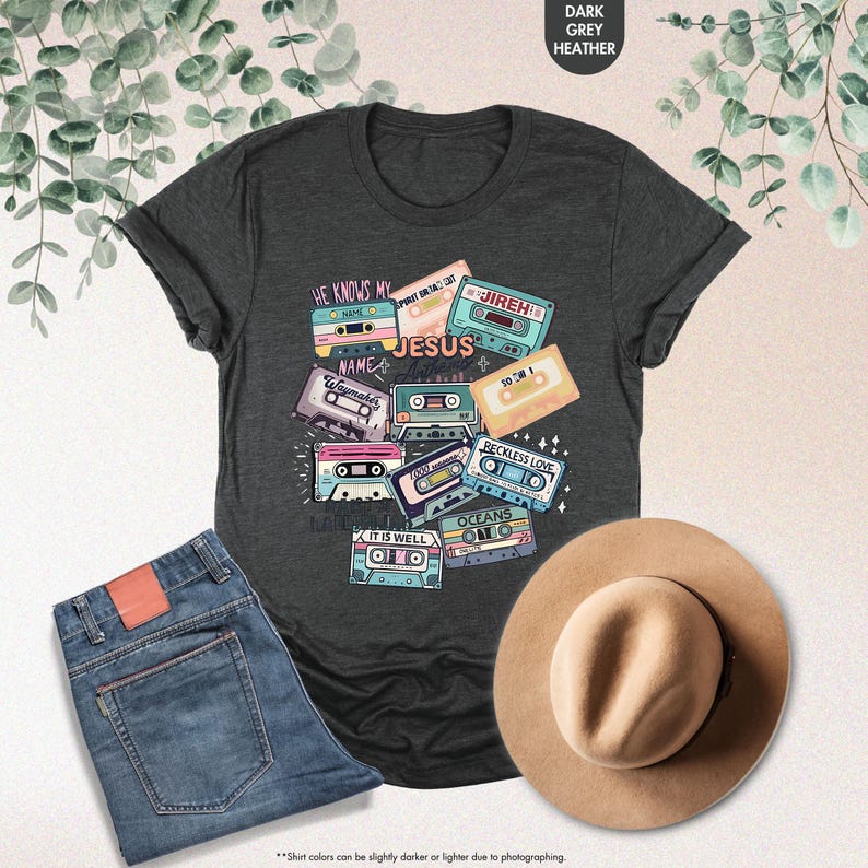 May include: A dark heather grey t-shirt with a design of vintage cassette tapes featuring Christian song titles like "He Knows My Name", "Waymaker", "Jireh", "It Is Well", "Reckless Love", "Oceans", and "So Will I".