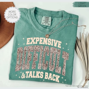 May include: A light green Comfort Colors t-shirt with the words "EXPENSIVE DIFFICULT & TALKS BACK" in a combination of pink and leopard print. A silver necklace with a pendant is draped across the shirt. The shirt is folded.